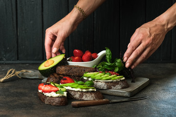 Toast or sandwich with avocado, cheese, strawberries, herbs and seeds on a dark background. Female hands serve a dish. An idea for bruschetta or for a healthy snack. Healthy vegan breakfast.