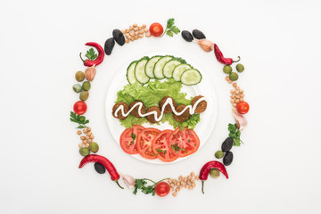 top view of vegetables arranged in round frame around falafel with sauce on plate on white background