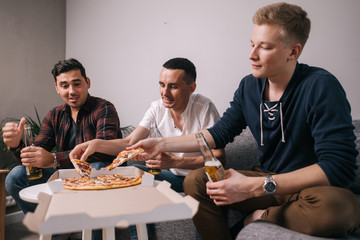 Group of young man eating pizza at the home. Guys are holding beer bottles in their hands.