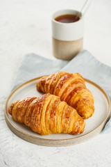 Two delicious croissants on plate and hot drink in mug. Morning French breakfast with fresh pastries. Light gray background, vertical