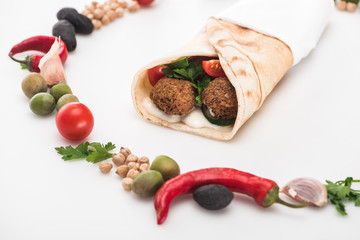 close up view of chickpea, garlic, cherry tomatoes, parsley, olives, chili pepper, green onion arranged in round frame around falafel in pita on white background