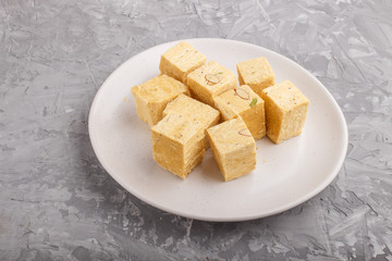 traditional indian candy soan papdi in white plate with almond and pistache on a gray concrete background. side view