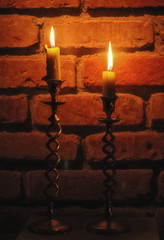 still life with two burning candles front view in vintage metal golden candlesticks on orange brick wall  background close up