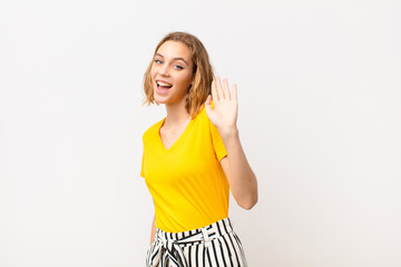 young blonde woman smiling happily and cheerfully, waving hand, welcoming and greeting you, or saying goodbye against flat color wall © kues1