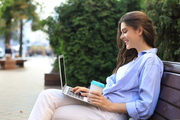 Beautiful woman sitting on bench holding coffee while using laptop outdoor.