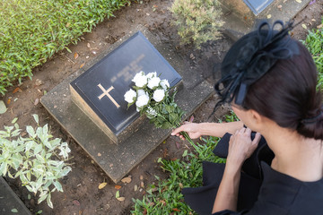 Mourning young woman laying white flowers on her family grave in beautiful green cemetery.