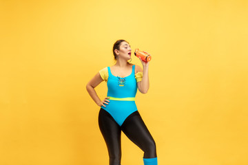 Fototapeta na wymiar Young caucasian plus size female model's training on yellow background. Stylish woman in bright clothes. Copyspace. Concept of sport, healthy lifestyle, body positive, fashion. Posing with bottle.