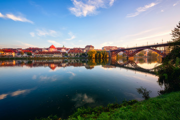 Fototapeta na wymiar Amazing view of Maribor Old city, Main bridge (Stari most) on the Drava river at sunrise, Slovenia. Scenic cityscape with blue sky and reflection, travel background for wallpaper or guide book