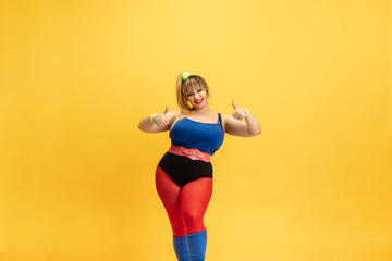 Fototapeta na wymiar Young caucasian plus size female model's training on yellow background. Stylish woman in bright clothes. Copyspace. Concept of sport, healthy lifestyle, body positive, fashion. Smiling with thumbs up.