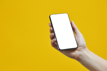 Fototapeta na wymiar Female hand holding black cellphone with white screen on a yellow background. Smartphone mockup with white screen