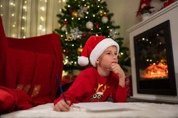 Fototapeta na wymiar the boy lies under the Christmas tree near the fireplace and writes a letter to Santa Claus