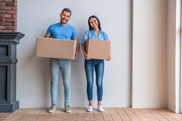Fototapeta na wymiar Couple holding moving boxes smiling at camera standing against wall in new apartment