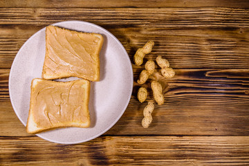 Glass jar with peanut butter and plate with sandwiches on a wooden table. Top view