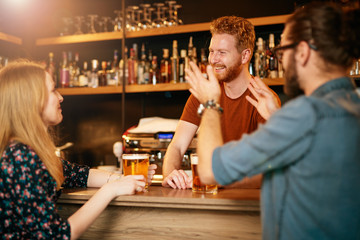 Cheerful friends leaning on bar counter, drinking beer and chatting with bartender. Night out.