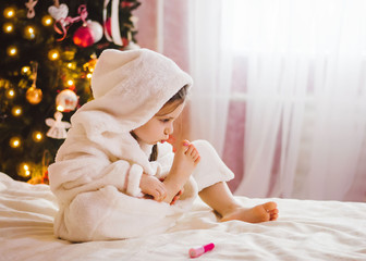 Fototapeta premium A little girl paints her nails. Girl sitting near the Christmas tree on the bed and paints her nails. Preparing to new year party