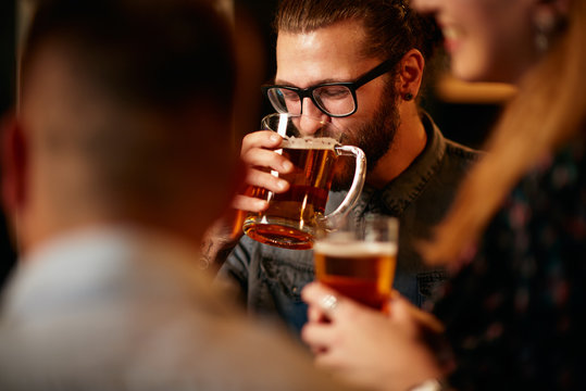 Handsome bearded caucasian young man with eyeglasses standing in pub with friends and drinking fresh beer.
