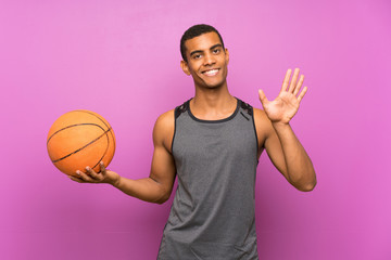 Young sport man with ball of basketball over isolated purple wall saluting with hand with happy...