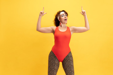 Young caucasian plus size female model's training on yellow background. Stylish woman in bright clothes. Copyspace. Concept of sport, healthy lifestyle, body positive, fashion. Smiling, pointing up.