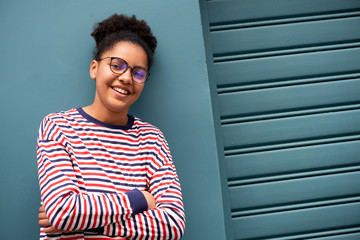 smiling african american teenage girl with glasses and arms crossed by blue wall