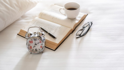 Alarm clock, book, cup of coffee and accessories on white bedding sheets with copy space. Morning lifestyle concept.