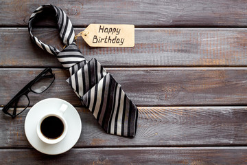 Man's birthday concept. Happy birthday text near male tie, glasses, coffee on dark wooden background top view copy space