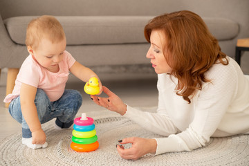 Little caucasian girl collects a pyramid with grandparents at living room. Grandmother plays with granddaughter on the floor