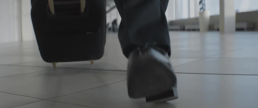 Unrecognizable man walking in black shoes and trousers with a luggage indoors, suitcase on wheels, airport or train station, business trip or travel concept, anamorphic dolly shot