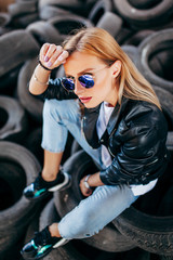 Portrait of fashionable blonde girl wearing a rock black style outdoors,  sits on old car tires in an abandoned factory