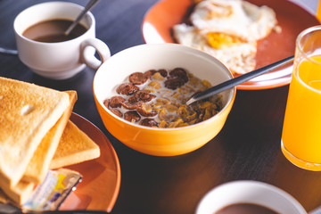 close up corn chocolate cereal with milk in orange blow with toasted, coffee, orange juice, and fried egg in background, with natural light, selective focus