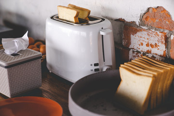 two slides of plain bread in white toaster, against rusty red brick wall, with pile of slices of...