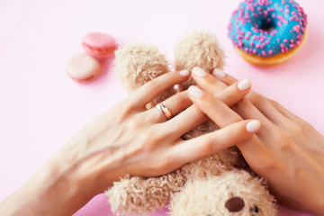 woman hands with manicure wearing wedding ring holdind teddy bear and macaroons on pink background