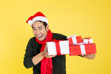 Happy Asian man wearing green sweater and Santa hat holding gift boxes on yellow background.