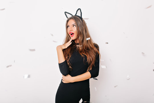 Curious dark-haired lady posing under shiny confetti. Indoor photo of good-humoured girl in black dress.