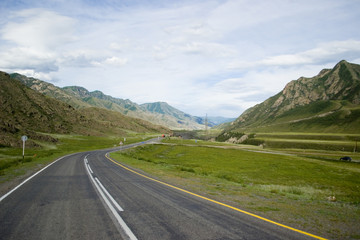 An asphalt road with a dividing strip, with road markings and signs runs along the mountains. The road is surrounded by green hills and mountains. Chuya highway in the valley of the Chuya River. Altai