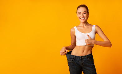 Girl In Oversize Jeans Gesturing Thumbs Up Smiling, Yellow Background - obrazy, fototapety, plakaty