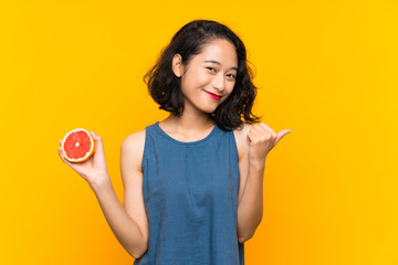 Young asian girl holding a grapefruit over isolated orange background pointing to the side to...