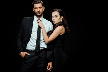 attractive girl in dress touching tie of boyfriend in suit standing with hand in pocket isolated on black
