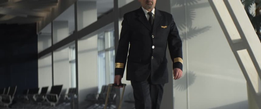 Aged flyer or pilot with suitcase with wheels walks through empty airport terminal in daytime. Elderly airman in a suit and cap walks down aboard the plane, anamorphic shot