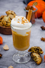  Pumpkin latte in a glass with spicy cream and a cinnamon stick on a gray stone background with cookies with chocolate chips. Autumn drink. Thanksgiving drink.