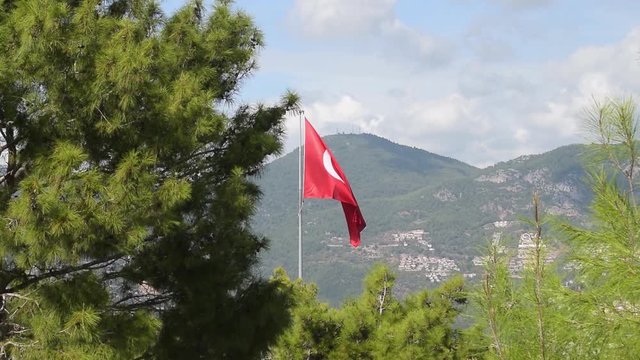 A large red flag flies in wind with conifers with mountains in the background