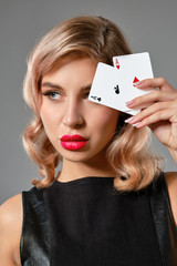 Blonde girl in black leather dress showing two playing cards, posing against gray background. Gambling entertainment, poker, casino. Close-up.