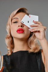 Blonde girl in black leather dress showing two playing cards, posing against gray background. Gambling entertainment, poker, casino. Close-up.