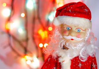 Toy, figurine, Santa Claus. Toy Santa Claus on the background of garland lights. Face, parts of the body of Santa Claus. New Year's toy in macro. The suit is red, a large gray white beard.