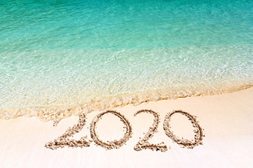 Happy New Year 2020, lettering on the beach with wave and clear blue sea. Numbers 2020 year on the sea shore, message handwritten in the golden sand on beautiful beach background. New Years concept.