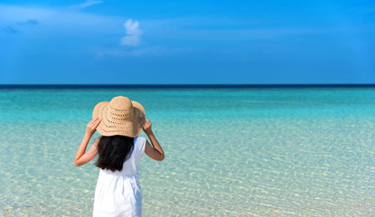 Fototapeta na wymiar Back of young Asian girl walking on white sand beach with clear blue sea and sky. Teenager girl wearing sun straw hat, white dress enjoy vacation. Outdoor summer vacation travel concept, copy space.