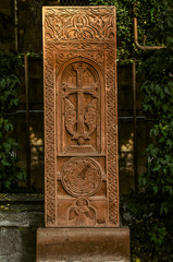 Fototapeta na wymiar A red tuff khachkar with a floral pattern over an arch with a cross,a figure of a Saint and a round symbol of eternity on the Alley of stone crosses in Gyumri