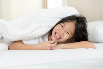 Portrait of happy young Asian girl lying on the tummy in bedroom after waking up and smiling under white blanket in the morning after good night sleep. Cheerful kid model with relaxing in the morning.