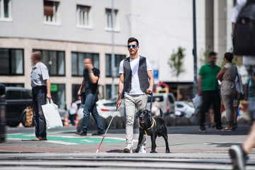 Young blind man with white cane and guide dog walking on pavement in city.