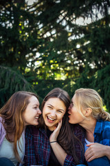 Three cheerful girls whisper and gossip against green foliage in the park. Women joke and laugh, vertical image.