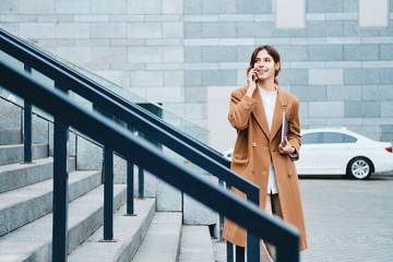 Young smiling casual businesswoman in coat with laptop happily talking on cellphone on city street